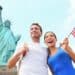 A Happy Tourist Couple, Holding A Small USA Flag With The Statue Of Liberty On The Background