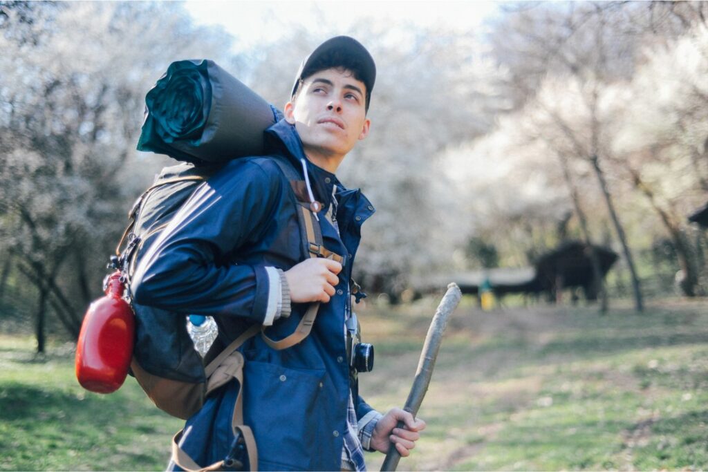 Male Wearing a Backpack Holding a Stick on a Solo Road Trip (Source: Canva)