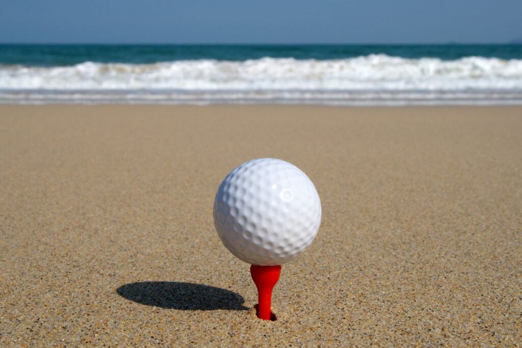 a golf ball lying on the beach, poised to be hit into the ocean